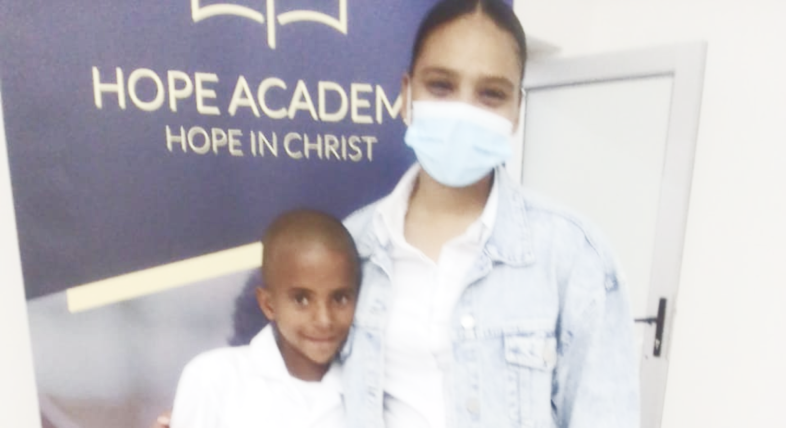 lady wearing mask standing next to little boy wearing school uniform